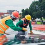 Two workers installing rooftop solar panels safely equipped with PPE and tools.